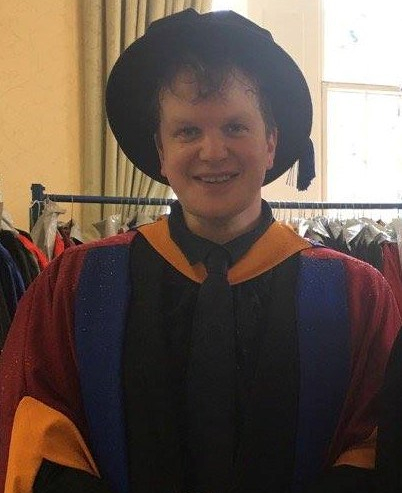 James, a white male with brown hair, dressed in colorful yellow, blue, red, and black graduation robes. He is wearing a black graduation hat.