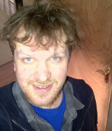 A selfie of James, a white man with blue eyes and a beard and mustache. His brown hair is windswept and he wears a blue T-shirt and black jacket. He looks pleased and relieved.