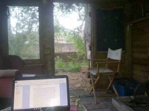 The inside of a rustic cabin, facing the open door to the outside. A computer sits open to James' book open in a Word document.