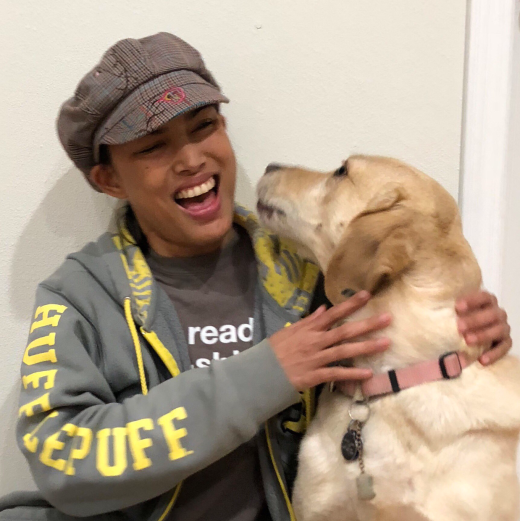 Elyana and her yellow lab guide dog. Yana is laughing with joy as her dog turns to lick her face. Yana wears a green Hufflepuff sweater and a plaid hat.
