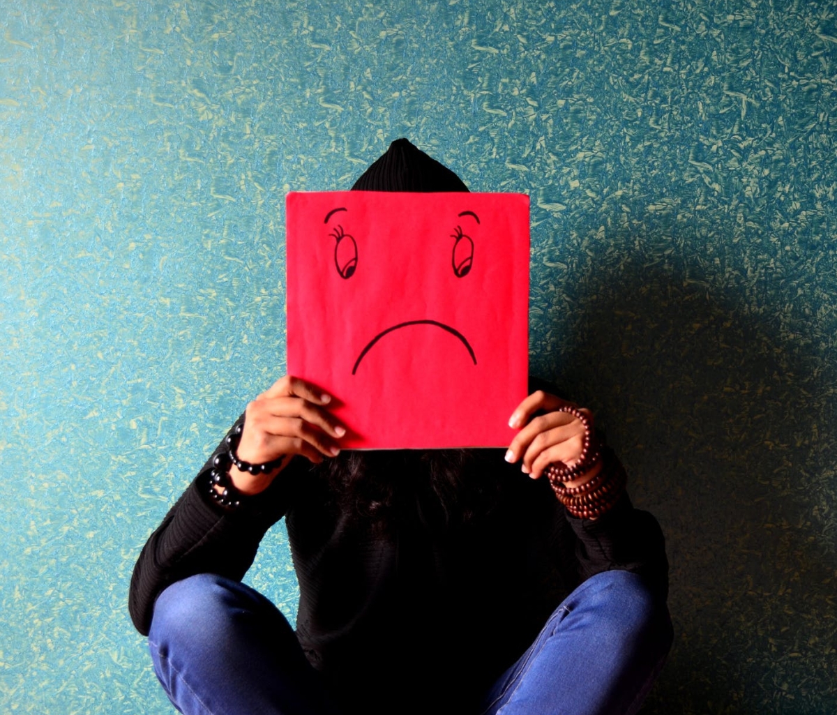 A picture of a person sitting cross-legged up against a wall. The person is holding a red square of paper up to cover their face; there is a frowning face with downcast eyes drawn on the paper.