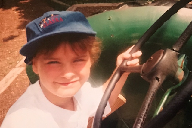 Alex as a young child, sitting on a play tractor, squinting into the sun and smiling faintly at the camera.